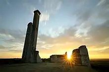 photo à contre-jour des ruines d'une construction en pierre. À gauche, une tour surmontée d'une tourelle carrée a gardé son aspect. À droite, les murs sont tronqués. Le soleil levant s'immisce dans une trouée des remparts.