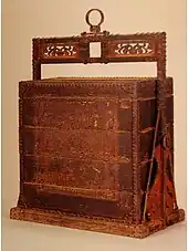 Coffre fermé à quatre étages et couvercle, maintenus par une sorte de cadre en bois dont la partie supérieure, ouvragée, est surmontée d’un anneau.