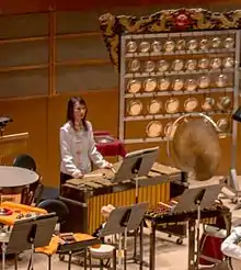 photo de la section percussion d'un orchestre avec une femme jouant d'un instrument au centre et derrière elle, à droite, un yunlao à 37 gongs.