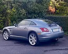 Une voiture grise, 2 portes, garée devant une haie et vue de l'arrière gauche.