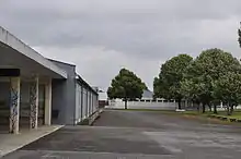 Autour d'une cour goudronnée, deux bâtiments techniques (aux toits d'usine) se font face. À droite, des bosquets d'arbre. À gauche, au premier plan, on distingue le porche d'une cour couverte.