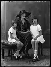 Une princesse portant une large capeline à plumes pose assise entre son fils (à gauche), vêtu de clair et sa fille portant une robe claire.