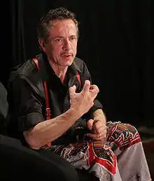 La photo montre Clive Barker parlant avec animation pendant une conférence. Il porte une chemise et un pantalon sombres et des bretelles rouges.