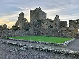 Photographie d'un cloître monastique dont il ne reste que les fondations. L'église mieux conservée est au second plan.