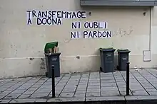 Sur un mur en extérieur, sont collés des feuilles avec des lettres noires peintes. Elles forment les phrases « Transfemmage à Doona » et « Justice pour Doona ».