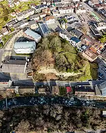 Colline entourée de bâtiments et coupée au bas de l'image par une falaise sous laquelle se trouvent une route et une rivière.