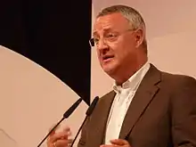 Photographie en couleur d'un homme. Il porte des lunettes. Devant lui se trouve deux microphones.