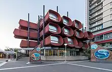 Une photographie du conservatoire de Montreuil prise en contre-plongée. Le bâtiment se distingue par son architecture originale, évocatrice du style des années 70 : des sortes de "cellules" tubulaires, de couleur rouille et dotées de vitres à leurs extrémités, constituent le bâtiment sur 3 étages.