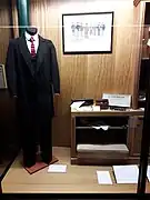 Photo d'une vitrine où est exposé un costume masculin de la fin du XIXe siècle. Il est formé d'un ensemble en drap de laine noir comprenant une redingote, un gilet et un pantalon, une chemise blanche ornée en coton d'un plastron, d'un col amovible et d'une cravate nouée en soie. Sur la droite, un support bas expose des accessoires tels qu'un coffret à boutons de manchettes et des gants blancs dans leur boîte.