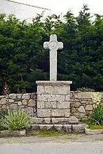 Vue d’une croix de chemin, avec pied en pierres de taille sur fond de haie de thuyas.