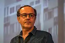 Homme cinquantenaire, aux cheveux bruns légèrement dégarnis sur le devant, portant une chemise sombre et des lunettes.