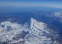 Un haut cône volcanique enneigé dominant des reliefs de type alpin plus modestes sous un ciel sans nuage, à peine brumeux à l'horizon.