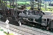Photo d'une maquette présentant au premier plan un signal et une voie ferrée ; au second plan, sous une structure métallique, deux locomotives à vapeur. La première locomotive fait 8 centimètres de haut, elle est vue depuis l'arrière ; elle est de couleur grise, délavée, montrant son usure. La seconde est cachée par la première, et semble être plus propre. Elle est verte à filets rouges. L'arrière plan présente une petite locomotive sur une autre voie parallèle, puis est fermé par une photographie de paysage.