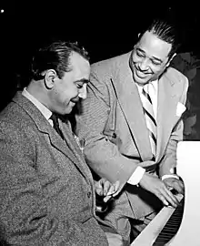 Deux hommes assis devant un piano blanc. Ellington a les mains sur le clavier, Django fume une cigarette