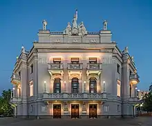 Un grand batiment blanc, façade avec trois portes, trois niveaux (deux étages), trois fenêtres à chaque étages, trois balcons ; couronnant la façade un groupe de statues.