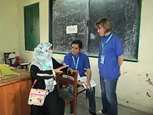 Photo montrant une femme assise voilée parlant à deux personnes, un homme et une femme habillés avec des tee-shits bleus
