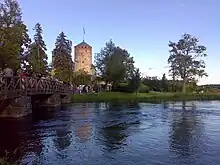 File d’attente sur un pont sur un petit lac ; en arrière-plan, un château entouré de pelouse, dont les tours sont visibles.