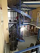 Photographie montrant l'escalier métallique peint en bleu-gris qui s'élève au centre du musée. La vue est prise à la hauteur du premier étage. En contrebas, on voit une ancienne machine industrielle exposée au rez-de-chaussée ainsi que le hall d'accueil avec les présentoirs de livres de la boutique.