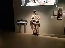 Une statue de cire de Louis de Funès en gendarme, à côté d'un panneau de signalisation indiquant la direction de Saint-Tropez.