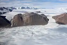 Petit glacier descendant d'un plateau cerné de sommets enneigés entre deux montagnes nues et brunes et confluant dans un large glacier en formant des crevasses concentriques.