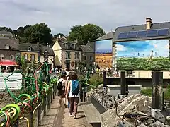 Photo d'un village entouré d'arbres. Sur un pont, des visiteurs franchissent un ruisseau. Sur les murs de maisons sont visibles des photos prenant la hauteur des bâtiments.