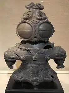 Photo couleur d'une statuette en terre cuite de couleur brun noir, sur fond beige.