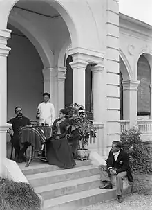 Photo représentant, sur le perron d'une habitation, un homme et une femme blancs, assis à une table. Un domestique asiatique se tient debout à leurs côtés. Un autre homme blanc est assis sur les marches de l'escalier.