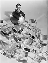 Photographie en noir et blanc d'un flacon de parfum en forme de boule couronnée d'un bouchon, le flacon se trouvant au milieu de petits cubes transparents.