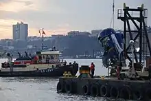 Photo de la récupération du moteur. À gauche, un navire supervise les opérations tandis qu'à droite, sur la berge, une grue tire le moteur endommagé du fleuve.