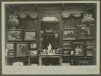 Vue d'une cimaise de la galerie Henry Vasnier sur laquelle sont accrochés des tableaux, d'autres sont posés à même le sol, contre elle. Au centre se trouve une cheminée surmontée d'un miroir dans lequel se reflète le photographe. Des sculptures et des objets d'art sont disposées sur sa tablette. D'autres sculptures sont posées sur des colonnes.