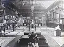 Vue d'ensemble d'une salle de la Galerie Vasnier avec la verrière comme source de lumière zénithale. Les murs sont recouverts de tableaux. Devant les cimaises, les sculptures sont disposées sur des sellettes et les objets d'art dans une vitrine. Des tapis recouvrent le sol.