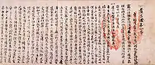 Photo couleur d'un texte écrit sur du papier en caractères chinois noirs sur fond rose, avec deux empreintes de mains en rouge à droite.