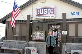 Une femme se tient devant l'entrée d'un bâtiment sur lequel il y a une pancarte «&nbsp;USO - Pat Tillman Memorial USO&nbsp;» et un drapeau américain.