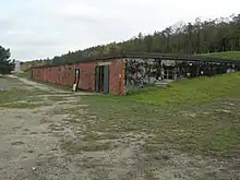 Ce long bâtiment d’un seul étage, au toit plat, semble être enfoncé dans la pente d’un terrain herbeux que clôture un bois de feuillus. La façade en briques est percée de deux portes et de six petites fenêtres.