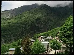 Photographie d'un village qui est enclavé dans sa forêt mais très ensoleillé ; on voit la chapelle au centre du village, entourée de maisons en lauzes et avec la rue arborée.