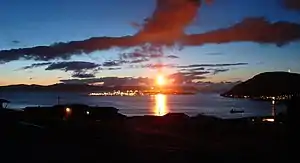 Au premier plan, des rochers noirs couvrent toute la partie inférieure de l'image. Une tour enflammée (la torchère) se dresse au loin et illumine une étendue d'eau et un ciel crépusculaire.