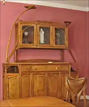 Photo en couleur d'un buffet en bois assez clair et poli avec quelques lignes arrondies, coin de table et dossier de chaise au premier plan