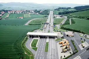 Vue aérienne d'une autoroute à quatre voie traversant les champs avec un petit village à l'arrière-gauche de l'autoroute. Au premier-plan se trouve une structure avec un toit blanc reposant sur quatre piliers blancs fins surplombant les quatre voies. À droite, on peut voir un parking avec des camions de couleur orange garés en diagonale.