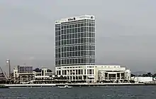 Photographie en couleurs représentant un bâtiment blanc aux nombreux étages, à la façade vitrée, sur le front de mer.