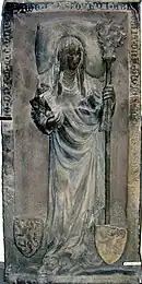 Photographie d'une épitaphe en relief montrant une femme qui tient une crosse et aux pieds de laquelle deux blasons sont posés.