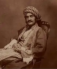 Photographie sépia d'un homme vêtu d'un habit du Moyen-Orient du XIXe siècle, portant une large moustache, assis sur une chaise avec les mains croisées sur ses jambes