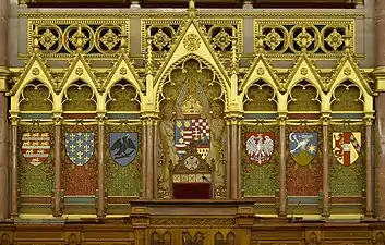 Derrière le pupitre du président de la Chambre des magnats sont représentés les blasons des principales familles ayant régné sur la Hongrie. Encadrant les armes du royaume, de gauche à droite : Arpadiens, Anjou, Hunyadi,  Jagellons, Zapolya, Habsbourg.