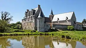 Photographie en couleur et panoramique d'un monastère fortifié et restauré, un plan d'eau entouré d'un tapis végétal visible au premier plan.