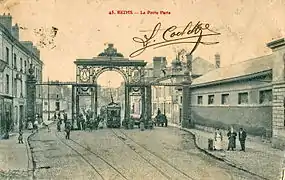Photographie d'une carte postale en noir et blanc représentant une largue rue barrée d'un porche en ferronnerie.