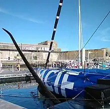 Vue de trois quarts arrière de l'avant d'un voilier bleu amarré au ponton. En premier plan, le foil bâbord.