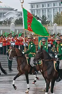 Cavalier sur un cheval en marche tenant un drapeau, suivi par deux autres soldats à cheval. En arrière-plan, une fanfare se tient devant les grilles d'un grand bâtiment blanc.