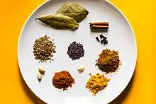 Plusieurs épices de différentes couleurs, entières ou en poudre, disposées en petits tas sur une assiette blanche.