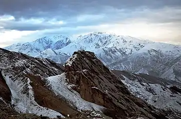 Une forteresse perchée sur un piton rocheux dans un environnement minéral et neigeux, avec de hautes montagnes en arrière-plan, ciel très couvert.