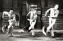 Trois hommes avec des couvercles de poubelles sur un pied chacun, en plein numéro de danse.