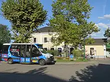 Photographie en couleurs d’un minibus bleu, blanc et noir stationné devant la gare ferroviaire de Rumilly, en arrière-plan.